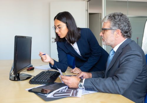 Focused male business leader and female assistant looking at statistic report on pc monitor, holding paper trading charts. Side view. Financial experts concept
